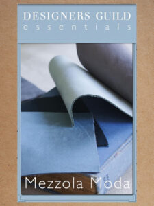 Foto mostrando a capa do catálogo da coleção de tecidos chamada Mezzola Moda da Designers Guild Essentials. A capa é azul claro com uma foto retangular com amostras de tecidos lisos em tons de azul sobre livro antigo fechado de capa azul.