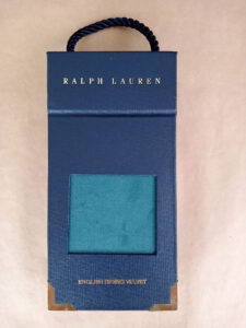 Foto do catálogo de amostras de tecidos. Capa dura, fundo azul, com um recorte retangular mostrando uma amostra azul de um dos tecidos. Com textos: Ralph Lauren, English Riding Velvet.