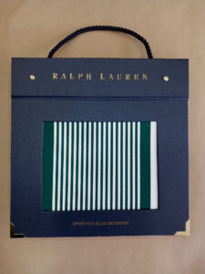 Foto do catálogo de amostras de tecidos. Capa dura, fundo azul, com um recorte retangular mostrando uma amostra listrada nas cores branco e verde de um dos tecidos. Com textos: Ralph Lauren, SPORTING CLUB OUTDOOR.