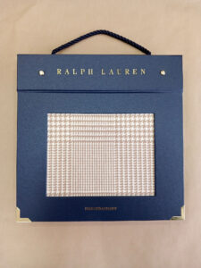 Foto do catálogo de amostras de tecidos. Capa dura, fundo azul, com um recorte retangular mostrando uma amostra estampada com padrões gráficos nas cores branco e bege de um dos tecidos. Com textos: Ralph Lauren, HABERDASHERY.