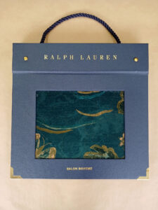 Foto do catálogo de amostras de tecidos. Capa dura, fundo azul, com um recorte retangular mostrando uma amostra estampada com cores azul, verde e dourado de um dos tecidos. Com textos: Ralph Lauren, SALON BOEME.