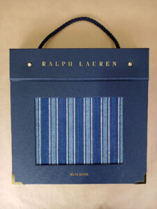 Foto do catálogo de amostras de tecidos. Capa dura, fundo azul, com um recorte retangular mostrando uma amostra listrada com cores azul e branco de um dos tecidos. Com textos: Ralph Lauren, BLUE BOOK.