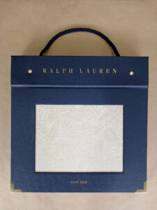 Foto do catálogo de amostras de tecidos. Capa dura, fundo azul, com um recorte retangular mostrando uma amostra lisa na cor branca de um dos tecidos. Com textos: Ralph Lauren, PARK ROW.