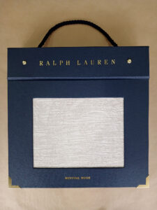 Foto do catálogo de amostras de tecidos. Capa dura, fundo azul, com um recorte retangular mostrando uma amostra lisa na cor branca de um dos tecidos. Com textos: Ralph Lauren, NEUTRAL BOOK.