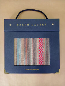 Foto do catálogo de amostras de tecidos. Capa dura, fundo azul, com um recorte retangular mostrando uma amostra listrada nas cores bege, azul e vermelho de um dos tecidos. Com textos: Ralph Lauren, ARCHIVAL TRAVELER.