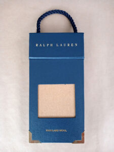 Foto do catálogo de amostras de tecidos. Capa dura, fundo azul, com um recorte retangular mostrando uma amostra lisa na cor bege de um dos tecidos. Com textos: Ralph Lauren, HIGHLAND WOOL.