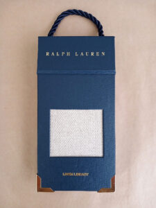 Foto do catálogo de amostras de tecidos. Capa dura, fundo azul, com um recorte retangular mostrando uma amostra texturizada na cor branca de um dos tecidos. Com textos: Ralph Lauren, LINEN LIBRARY.